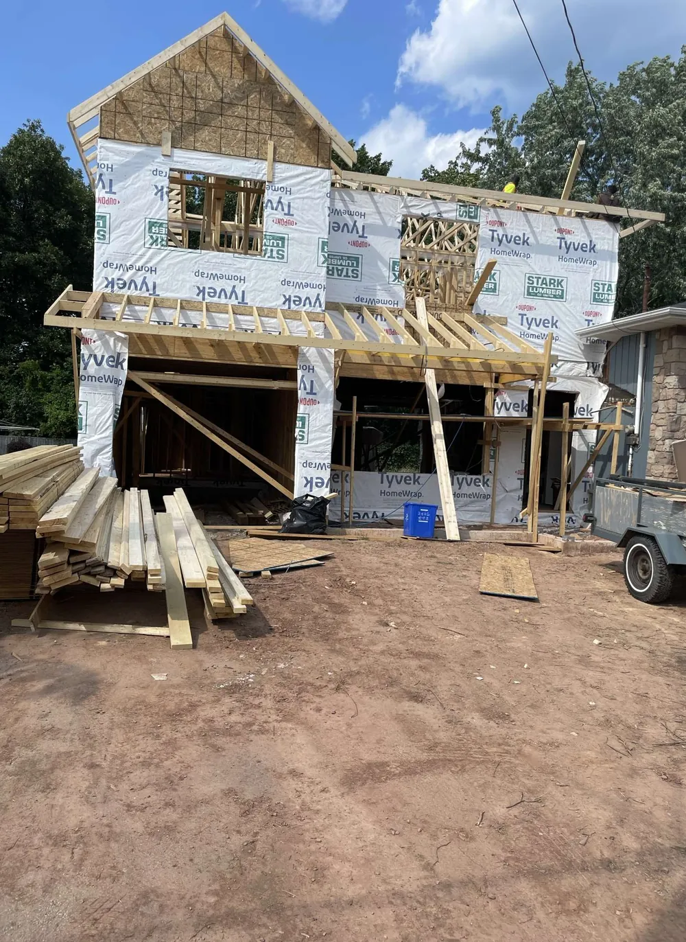 Two-storey house under construction, wrapped in house wrap with new roof framing and lumber stacked in the yard