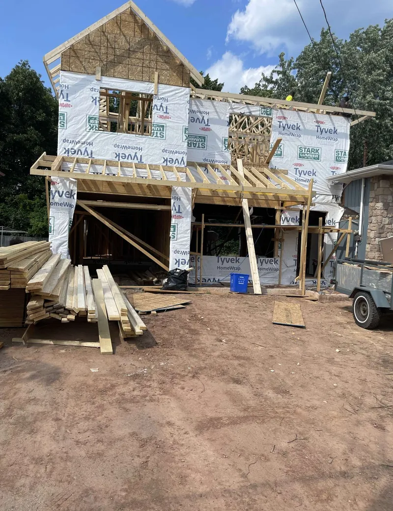 Two-storey house under construction, wrapped in house wrap with new roof framing and lumber stacked in the yard